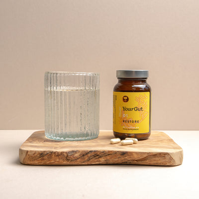 A ribbed glass of water, a brown bottle labeled "01 Restore" synbiotic supplement, and four white capsules are arranged on a wooden board against a neutral background to promote gut health.