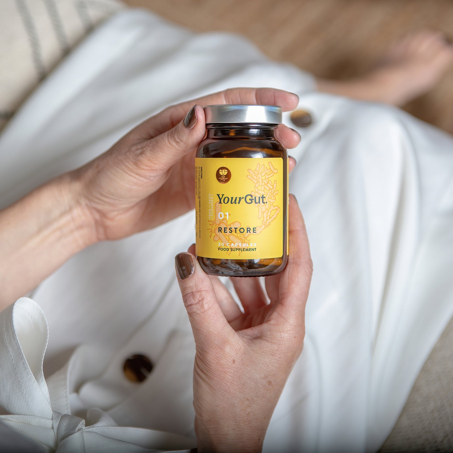 A person in a white dress holds a brown probiotic glass jar of 01 Restore, a synbiotic gut health ethical supplement with a yellow label and silver lid. Their nails are painted brown.