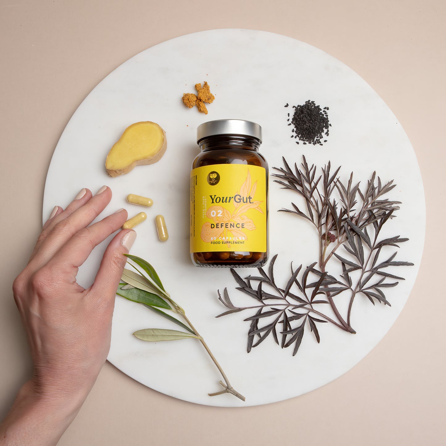A hand reaches toward a bottle of 02 Defence, an immune supplement for gut balance, on a white marble surface with ginger, propolis, capsules, black elderberry, olive leaf and black cumin seeds.