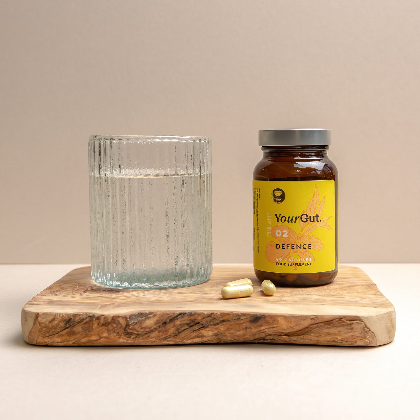 A glass of water, a brown bottle labeled 02 Defence, and three yellow capsules—an immune supplement for gut balance—are arranged on a wooden board against a beige background.